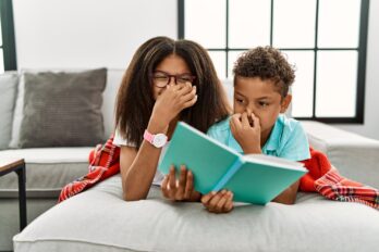 Two,Siblings,Lying,On,The,Sofa,Reading,A,Book,Smelling